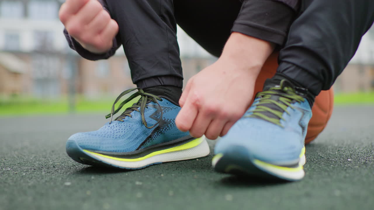 Corridore che si allaccia le scarpe da ginnastica sul campo, mani che annodano scarpe blu brillante accanto al pallone da basket, primo piano ad angolo basso, texture di asfalto, riscaldamento pre-partita mirato, giacca e pantaloni da jogging casual, luce nuvolosa, urbano