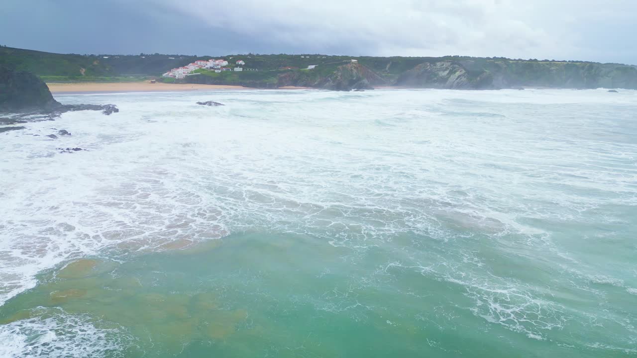 Waves crash on beach near Aljezur, Portugal, with cliffs and coastal village behind