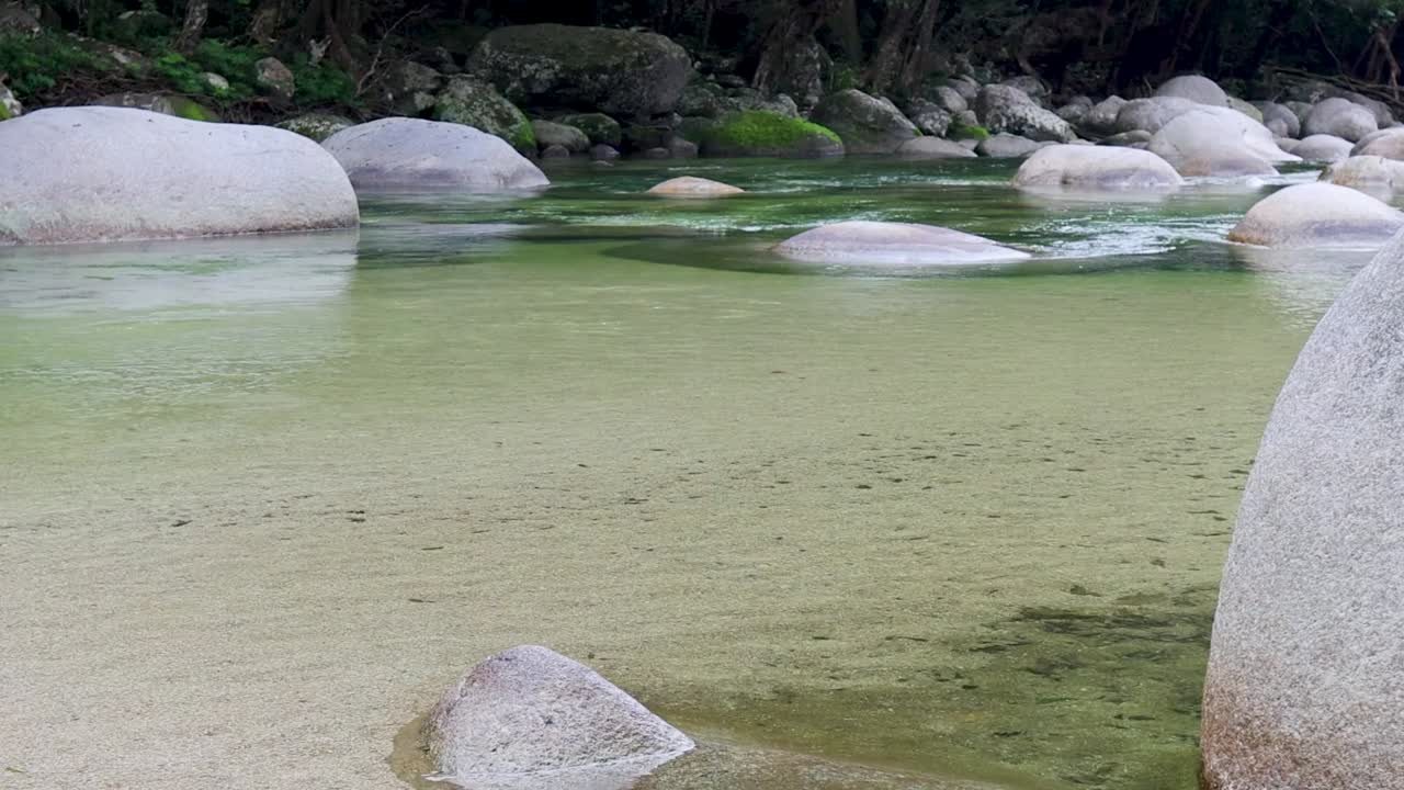 Gentle ripples move across a shallow, clear riverbed surrounded by large smooth granite rocks