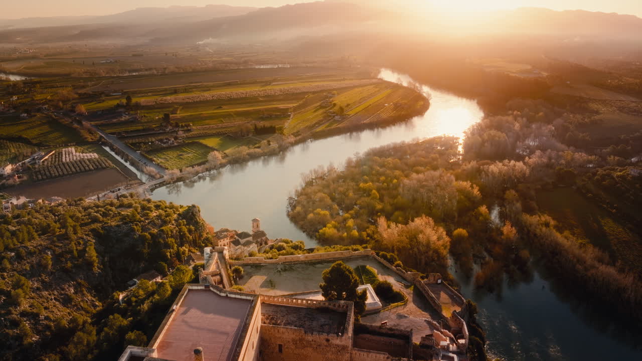 Ripresa aerea cinematografica 4K di un fiume al tramonto, con il Castello di Miravet, Spagna. Storico villaggio medievale sulle rive del fiume con torre della chiesa, tetti in tegole e un corso d'acqua serpeggiante. Luce dell'ora d'oro che si estende sulla campagna.
