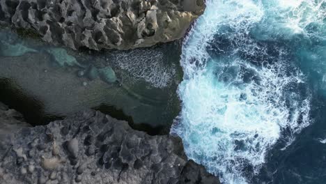 Dynamic-blue-ocean-waves-crash-into-shallow-rocky-inlet-in-Bali-aerial