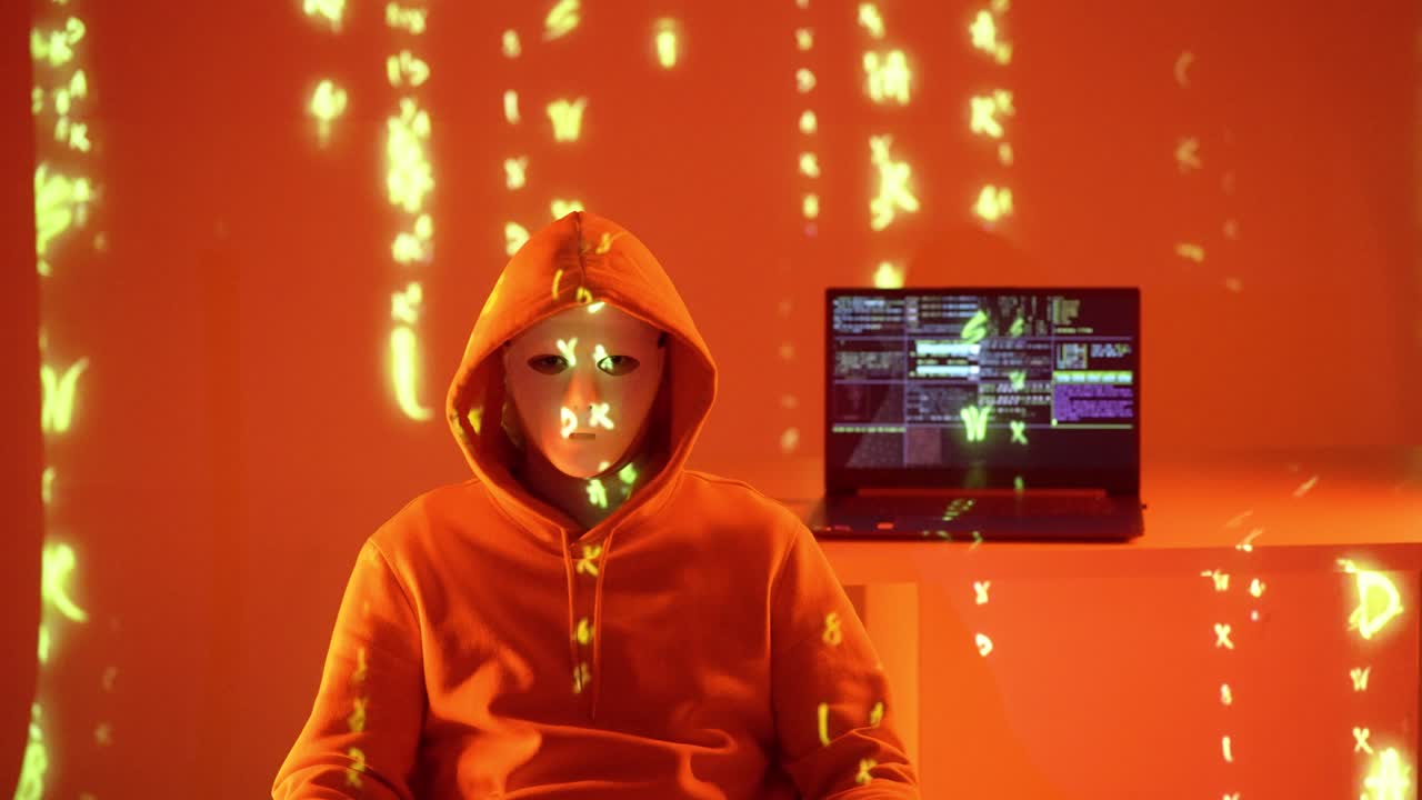 A mysterious hacker in an orange hoodie and white mask sits in a violet neon room with code projected around. A terminal-filled laptop glows in the back, visualizing secrecy and cyber tech