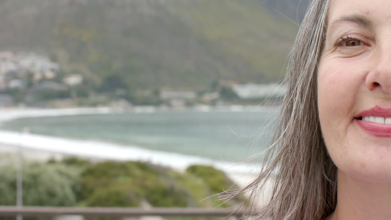 Senior woman smiling by balcony railing with breeze lifting hair, overlooking shoreline copy space