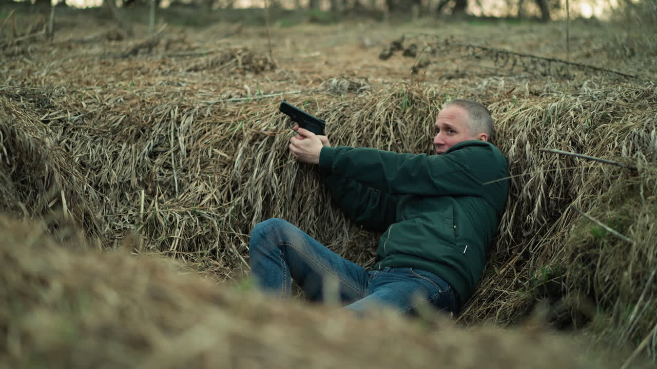 청바지와 검은 신발을 입은 사람이 건조한 풀을 가로질러 달리고 은 길에 누워 총으로 겨냥하는 동안 긴장되어 보입니다.