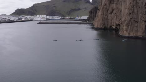 kayakers crossing bay near harbor of vestmannaeyjar with rocky cliffs, aerial