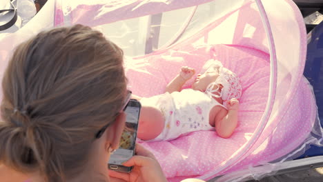 Baby-girl-in-pink-bassinet-and-mother-browsing-web-on-cell-outdoor