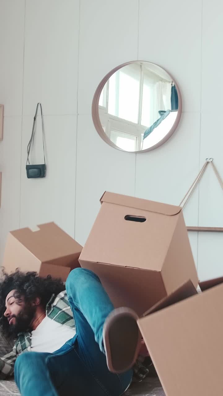 Young positive casual arabian man with empty moving boxes falls on sofa