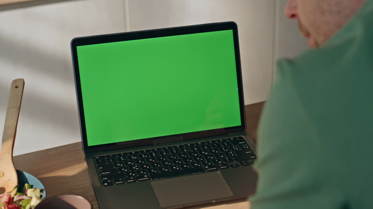 Primo piano di mani di un uomo sconosciuto che digitano su un laptop con schermo verde in cucina. Computer con chromakey.