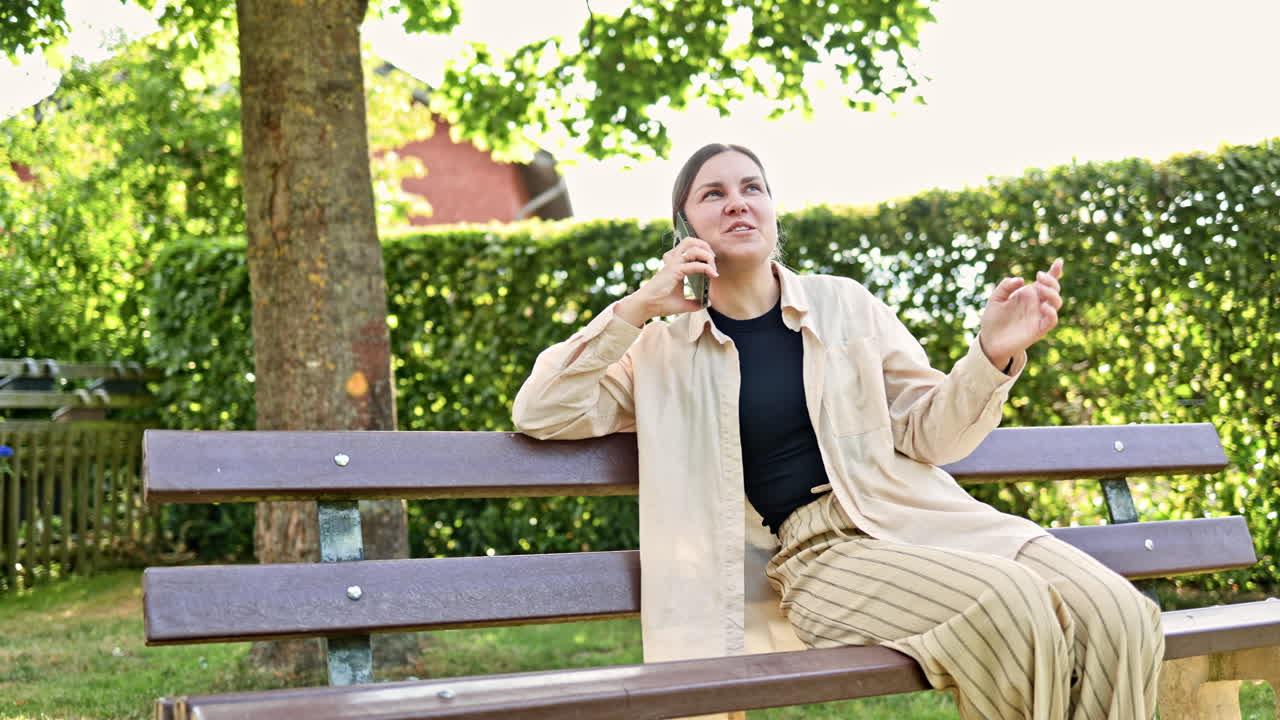 Una donna allegra si gode felicemente una giornata di sole mentre è seduta su una panchina del parco e parla animatamente al suo smartphone, mostrando un'atmosfera rilassata all'aperto, sorrisi genuini e un'aria spensierata.