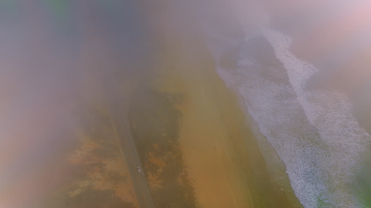 Foamy waves coming to the sandy shore. Drone descending through the fog over the road going by the beach.
