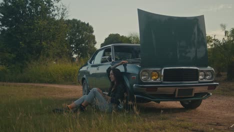 woman sitting beside broken-down vintage car on a country road