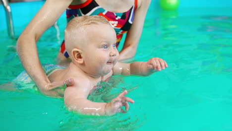 lovely blond baby boy wearing a diaper learning to swim. unrecognized coach holding a kid in the swimming pool.