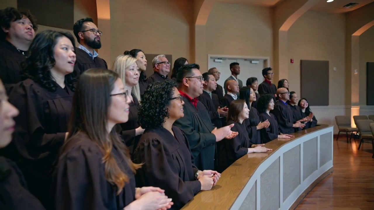 Reagendo al segnale del direttore, un coro in abiti scuri canta e applaude in una cappella vicino a una ringhiera curva