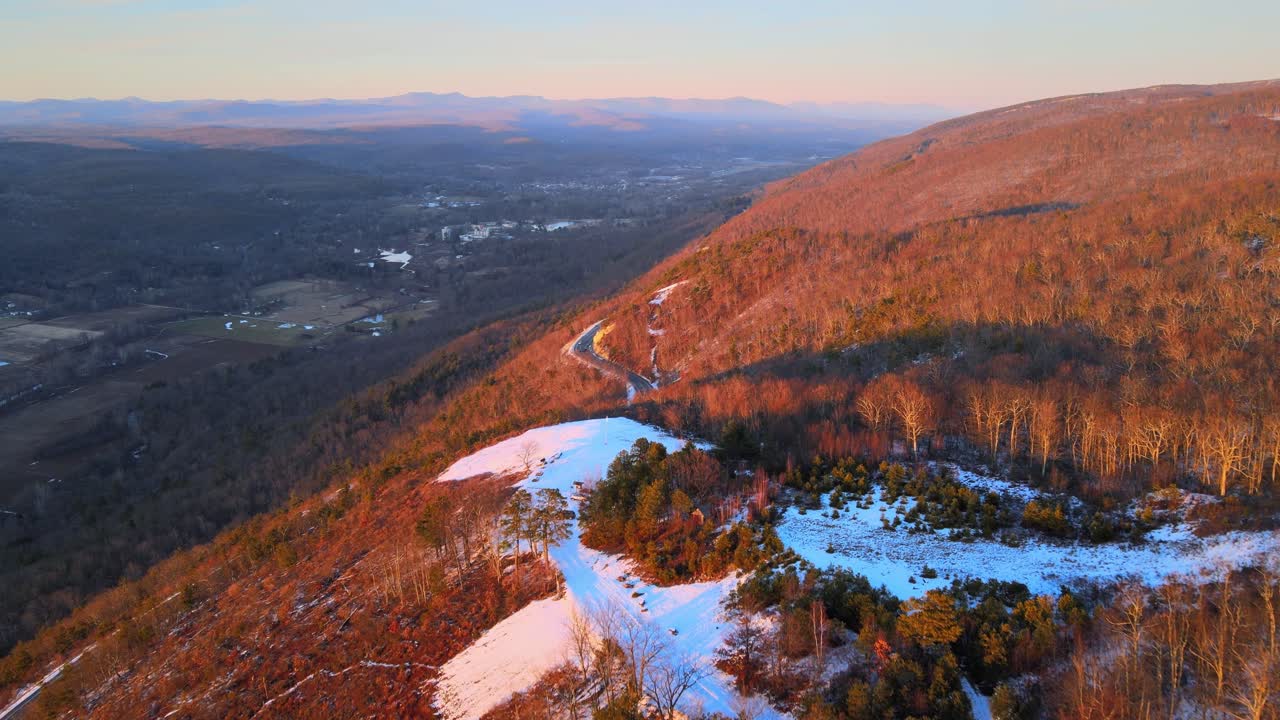 volando alto sobre una montaña con una ligera capa de nieve y una carretera escénica con un valle grande y expansivo debajo con montañas a lo lejos durante la puesta de sol