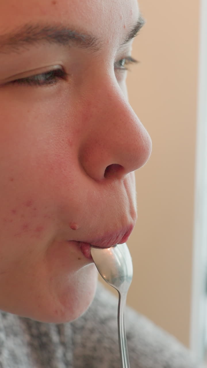 primer plano de un adolescente blanco probando una cuchara en una cocina tranquila, luz natural suave, expresión contemplativa, sorbo suave, acné visible en la mejilla, cuchara y mano de metal, momento doméstico íntimo, aspirante a catador