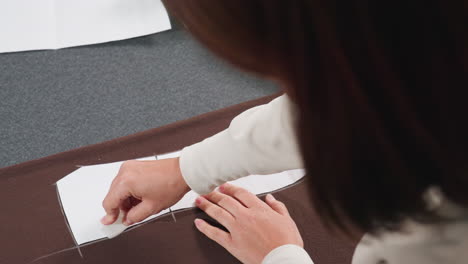 elegant woman with short brown hair making precise markings on white paper pattern placed over dark brown fabric in sewing workspace, carefully sketching outlines for garment design preparation