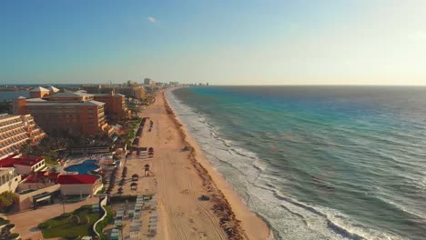 Las-Olas-Del-Mar-Tropical-Se-Estrellan-En-La-Costa-De-Arena-Blanca-Con-Un-Cielo-Azul,-Los-Rayos-Dorados-Del-Sol-Brillan-En-El-Mar-Turquesa-Junto-A-Los-Resorts-De-La-Zona-Hotelera---Laguna-Nichupté-En-Cancun,-México---Drone-Panning