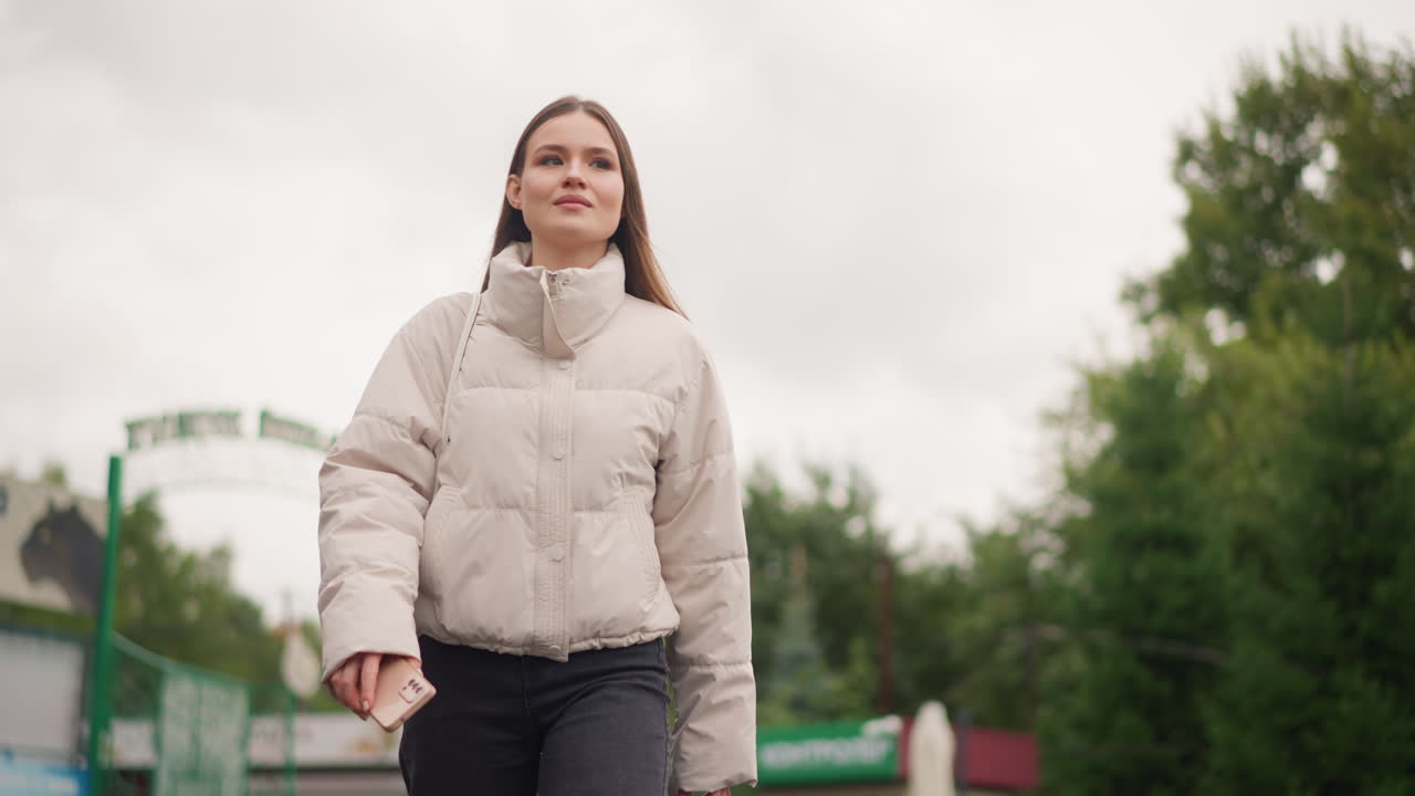 Una mujer joven caminando por la entrada de un parque, cielo nublado, chaqueta acolchada color crema, manos relajadas, paso firme junto a una verja de hierro verde y señalización, árboles enmarcando el fondo, natural y confiada