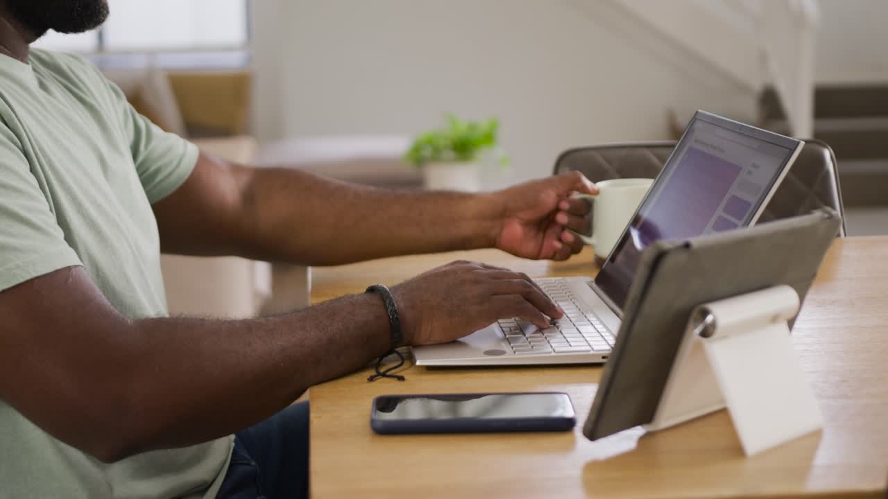 African mid-adult man starting work at wooden desk, typing on laptop and sipping from white mug