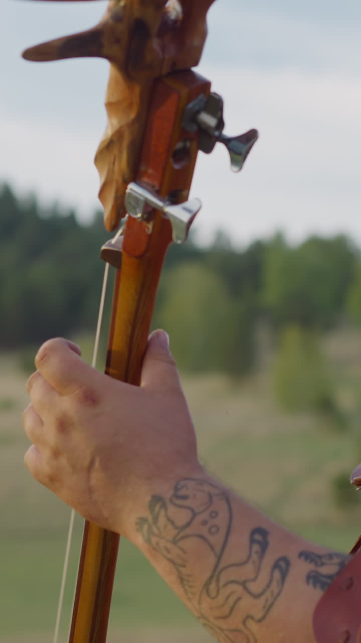Musician with headband and fur cover holds tovshuur sitting in valley close side view. Art and cultural heritage of Siberian folks. Music instrument