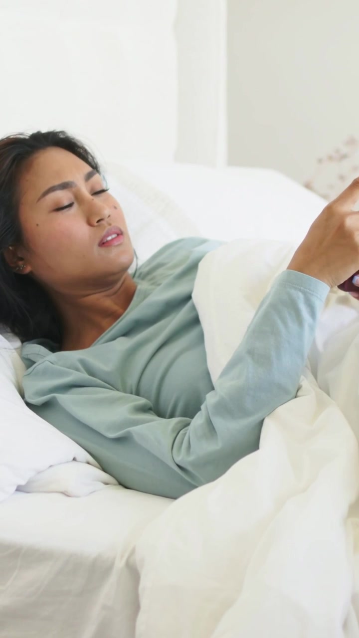 Video verticale: Donna in top blu che guarda il telefono viola a letto, tirando il piumone per dormire.
