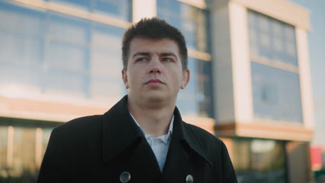 hombre de traje negro ajustando el cuello con una expresión enfocada al aire libre, el fondo destaca edificios modernos de vidrio y estructuras urbanas