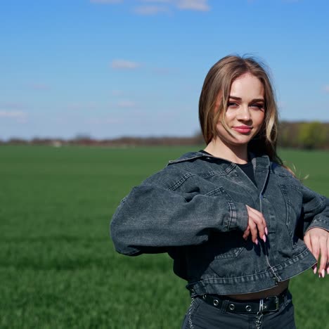 Attractive girl posing on camera outdoors. Portrait of a young woman with long hair in denim jacket looking on camera on green field background