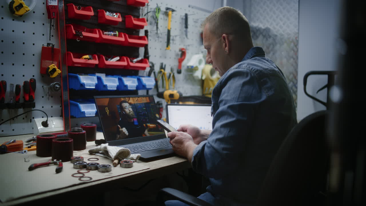 Worker Watching Online Trading Education on Laptop, Monitoring Real-Time Stocks and Exchange Market Charts Using Smartphone. Man Remote Learning Trading During Working Day in Workshop. Vertical Shot.