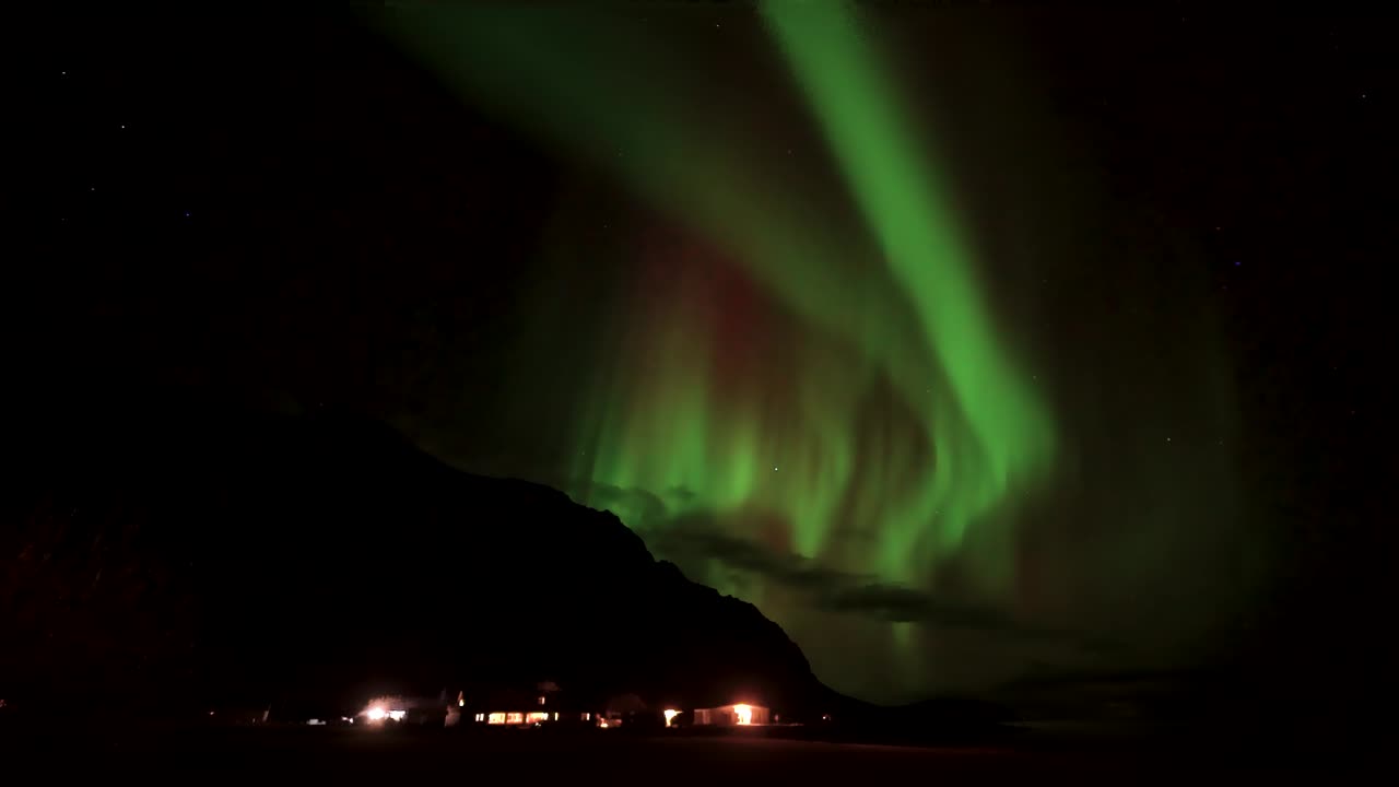 Green Aurora Borealis Swirling Above Dark Mountain Silhouette At Night In Iceland. Illuminated Houses At Base. timelapse