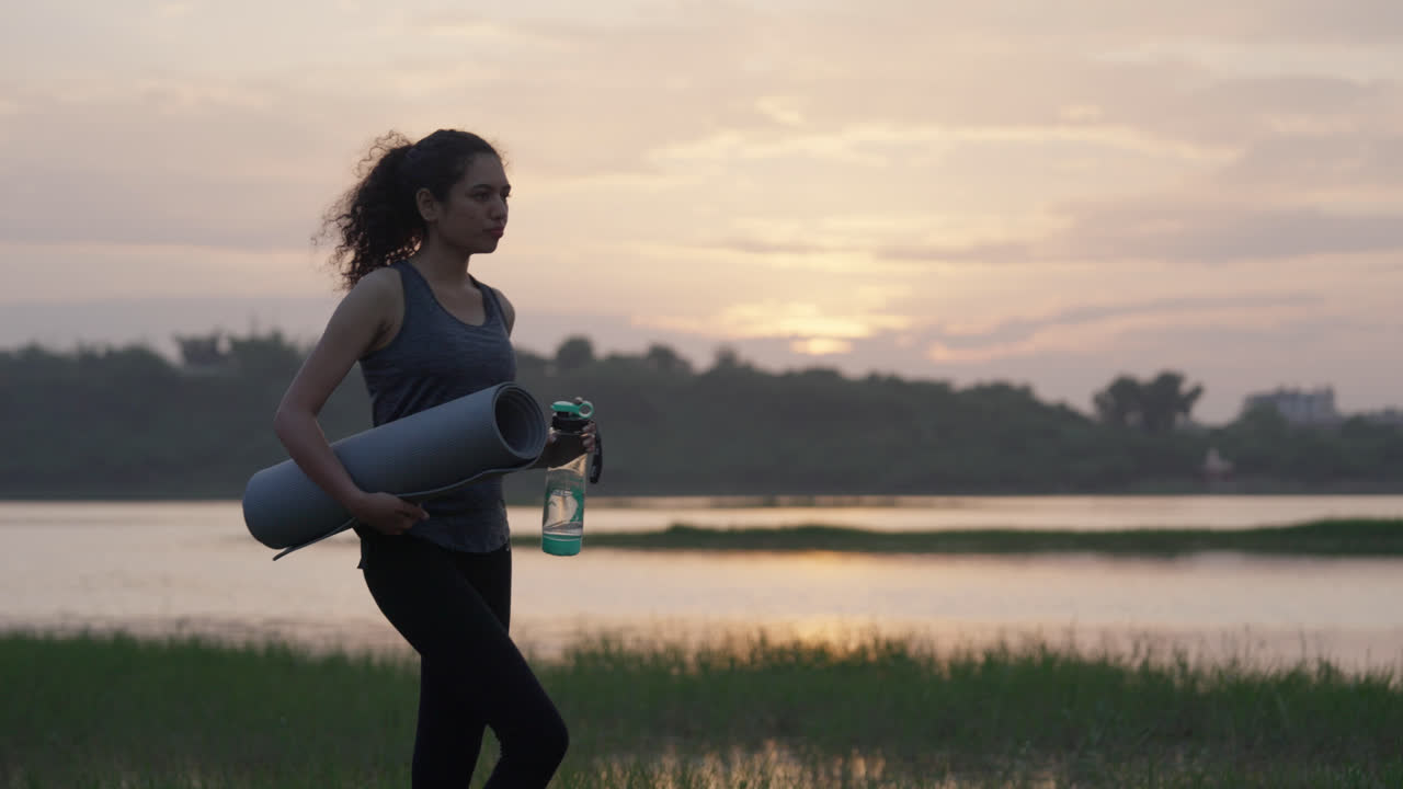 A South Asian woman holding her yoga mat and water bottle beside a misty lake, the soft sunrise light spreading across the horizon