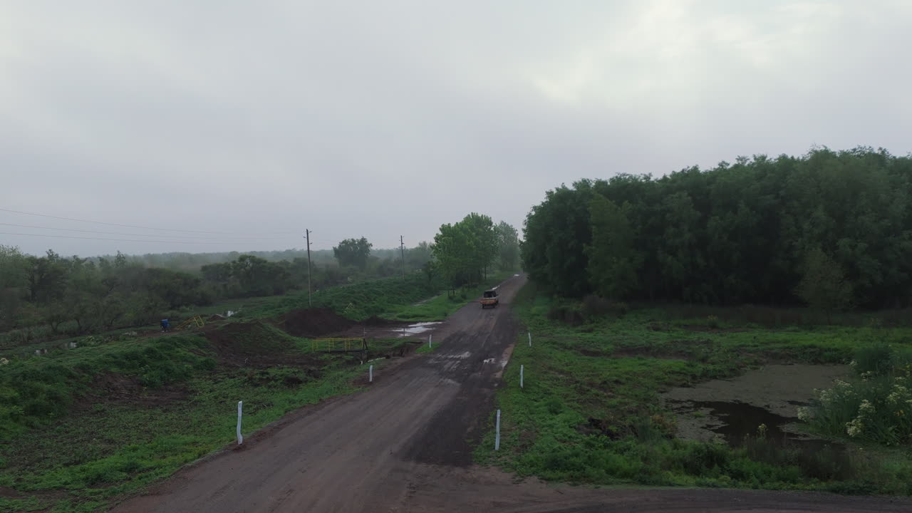Rural dirt road curving through a dense green forest with moving old car on an overcast weather.