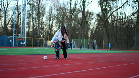 little kid follows his mom kicking a ball at the stadium. mother and baby playing football cheerfully.