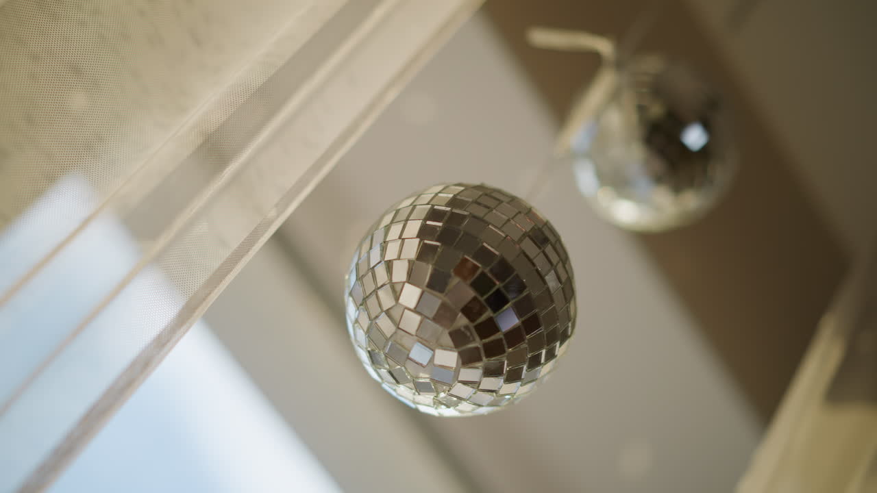 Closeup shiny disco ball decoration hanging near window with second blurred ball in background, reflecting light on mirrored surface creating festive stylish modern interior atmosphere with sparkling details