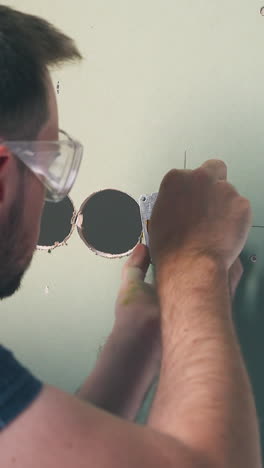 cu, dolly shot: electrician in protective glasses and blue shirt repairs plug on wall with screwdriver in room close back view