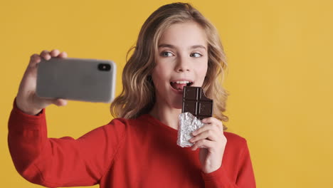 teenage caucasian girl eating chocolate bar and taking selfies with smartphone.