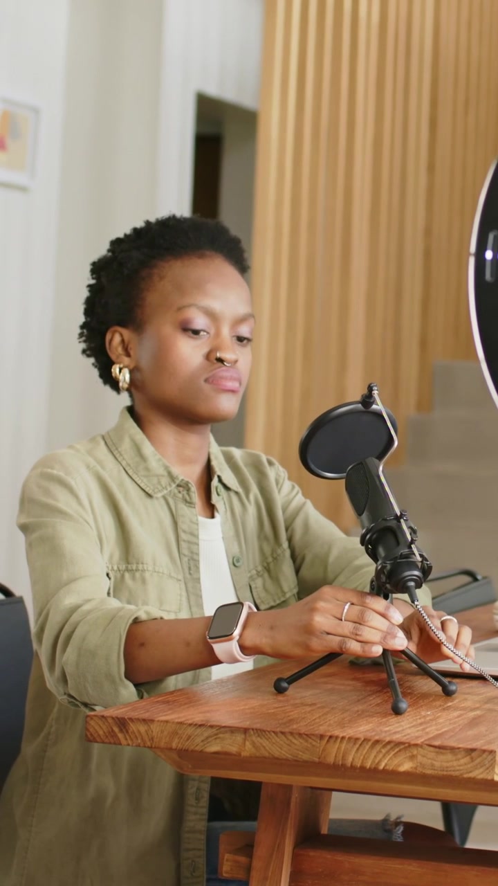 Vertical video: Checking smartwatch, woman in olive shirt adjusting mic at home table near laptop