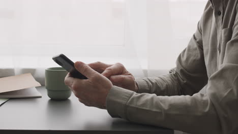 camera focuses on senior man hands using smartphone sitting at desk at home