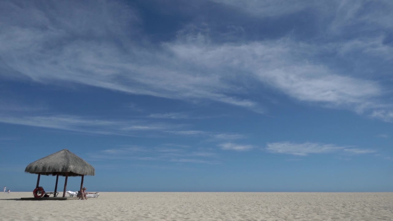 Ampia vista costiera con spiaggia sabbiosa, riparo con ombrellone di paglia e cielo blu drammatico con nuvole sottili sopra un'atmosfera pacifica del litorale.