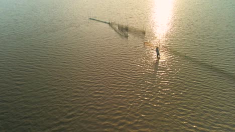 Pescador-Pescando-Con-Red-En-La-Vasta-Y-Expansiva-Vía-Fluvial-De-Tonle-Sap,-Asia,-Vista-Aérea-De-Pájaro-Del-Pescador-Recortado-Lanzando-Su-Red-Durante-La-Puesta-Del-Sol-Dorado-Al-Final-De-La-Tarde,-Cámara-Lenta,-Espacio-De-Copia