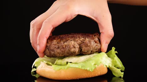 a hand carefully places a cooked beef patty onto iceberg lettuce and a burger bun against a black background, with even studio lighting and smooth camera movement