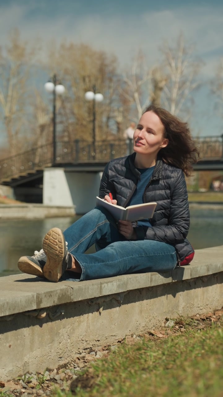 Una donna legge e scrive su un quaderno vicino all'acqua con un ponte, alberi e un prato sullo sfondo. La donna indossa una giacca scura sopra una camicia blu mentre tiene un libro aperto con una penna rossa visibile sulle pagine. Parco all'aperto.