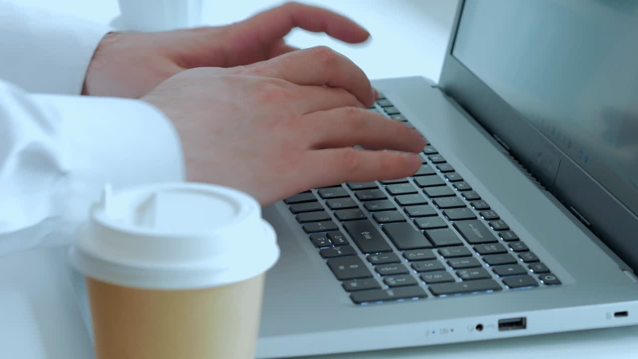las manos de los hombres presionan botones en un teclado de portátil, una taza de papel con una tapa de plástico en una mesa blanca