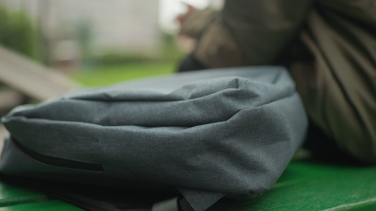 Close up of schoolbag resting on green bench outdoors with kid seated nearby adjusting posture while playing handheld game, blurred greenery and casual background