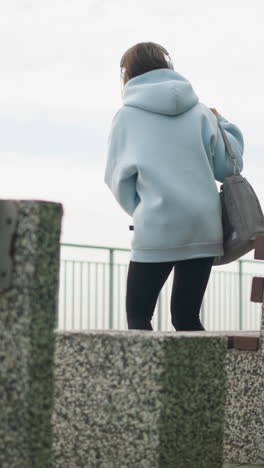 back view of young woman walking toward concrete bench to sit, carrying bag in peaceful park setting, with green leaves and railing in background, perfect for leisure, walking, and nature shots