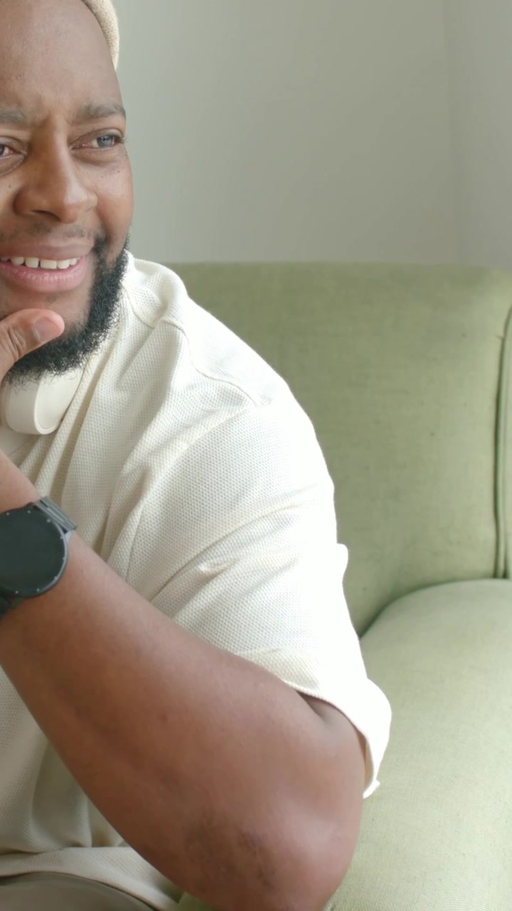 Video verticale: Un uomo con berretto a cui svanisce il sorriso sposta lo sguardo e si tocca il mento a casa, con uno smartwatch.