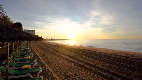 Kardanische-Aufnahme,-Die-Sich-An-Einem-Strohstrandschirm-Vorbeibewegt,-Um-Einen-Wunderschönen-Sonnenaufgang-Am-Strand-Von-Marbella-Zu-Enthüllen