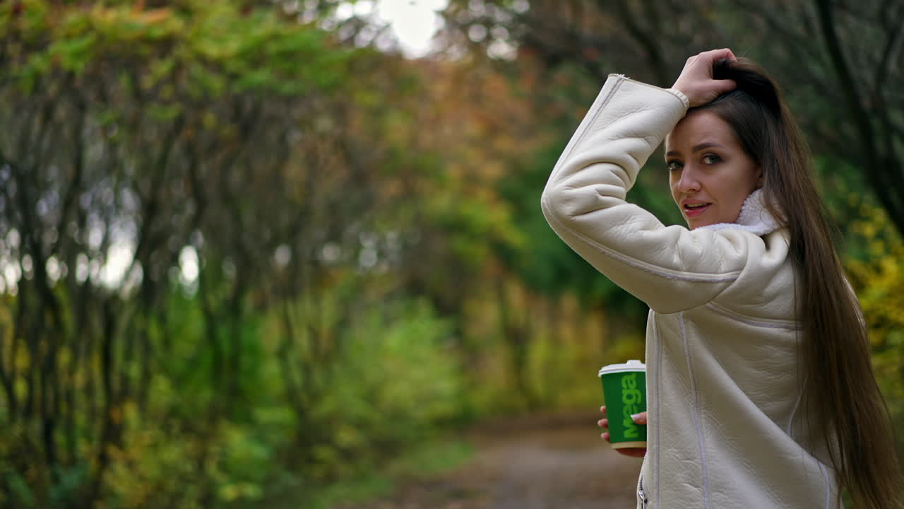 Attractive brunette lady touching her long hair walking in the park. Woman in warm jacket drinking coffee in the nature in autumn.