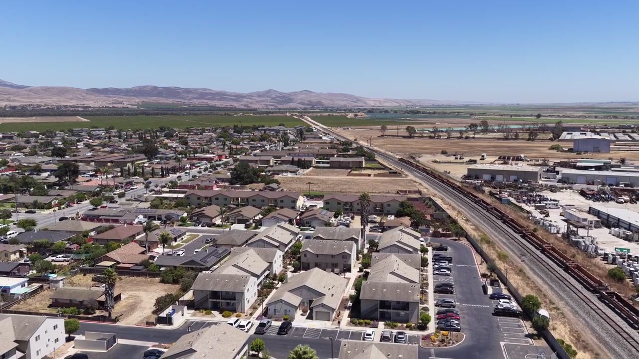 Aerial perspective flying over the residential neighborhoods and railroad tracks of Soledad, California, with distant agricultural fields and mountains creating a picturesque landscape