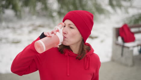 lady in red beanie and hoodie sipping water from pink bottle outdoors, blurred background showing bag on frosted bench, greenery, and iron rail in serene winter setting