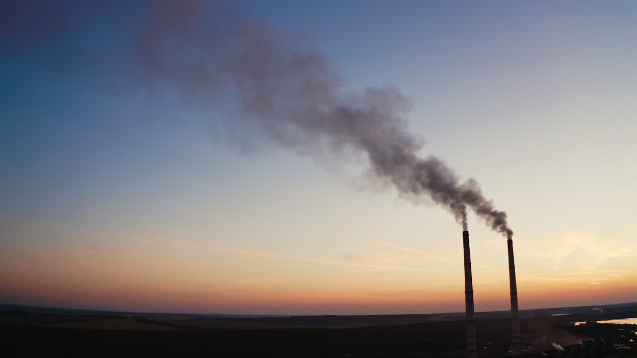 Factory with pipes among nature in the evening. Two industrial pipes with dark smoke pollute the environment at sunset.